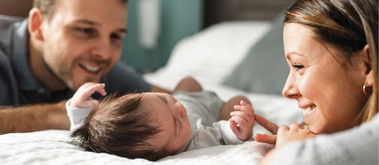 Mom and dad smile at their newborn baby lying on a bed between them.