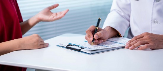 Clinician signs a form on a clipboard at a table as a patient sits nearby and gestures