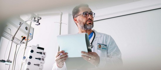 Male doctor with glasses and a beard holding a clipboard/folder in a clinical setting with medical equipment nearby.