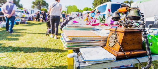 Outdoor flea market scene with tables holding stacks of magazines and a vintage rotary telephone on a wooden stand, shoppers in the background.