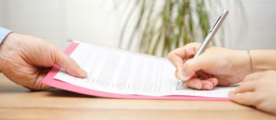 Two people signing a document on a pink folder at a wooden desk.