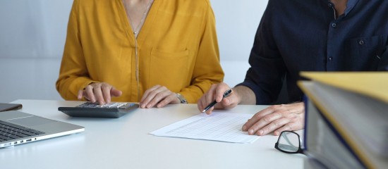 Two professionals reviewing a document at a white table, with a laptop, calculator, pen, and glasses nearby.