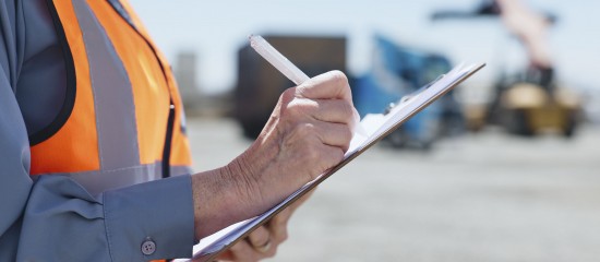 Construction worker in an orange safety vest writing on a clipboard at an outdoor worksite.
