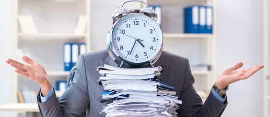 Overwhelmed office worker with a large alarm clock covering their head, surrounded by a tall stack of papers in an office setting.