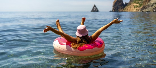 Person relaxing on a pink inflatable inner tube in calm blue sea, arms outstretched, wearing a pink sunhat, with rocky coastline in the distance.