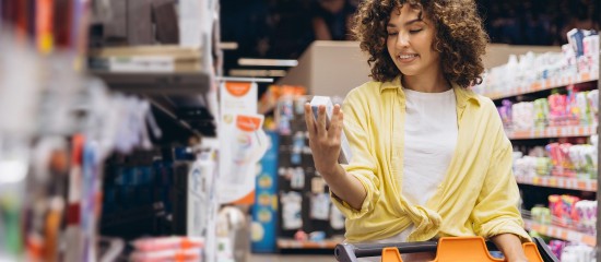 Young woman in a yellow shirt stands in a grocery store aisle, examining a product while pushing an orange cart.
