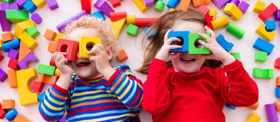 Two smiling children lie on a colorful floor, holding bright toy blocks over their eyes while surrounded by scattered blocks.