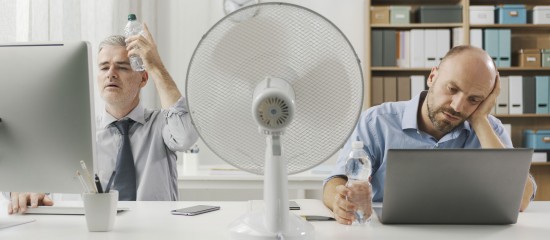 Two office workers sit at a cluttered desk with a large fan between them; one drinks water while the other rests his head on his hand near a laptop, looking stressed or tired.