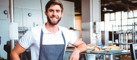 Smiling bakery worker in a striped apron standing beside a glass display with pastries behind him.