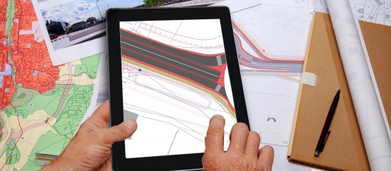 Hands holding a tablet that displays a construction/engineering map with red and black lines, surrounded by maps and blueprints on a desk.