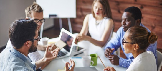 Five coworkers around a table in a modern office, collaborating with laptops and coffee cups on the table.
