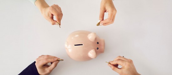 Four people are dropping coins into a pink piggy bank on a white surface, symbolizing shared saving.