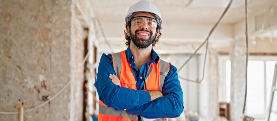 Construction worker in a hard hat and orange safety vest stands with arms crossed inside a partially built room, smiling at the camera.