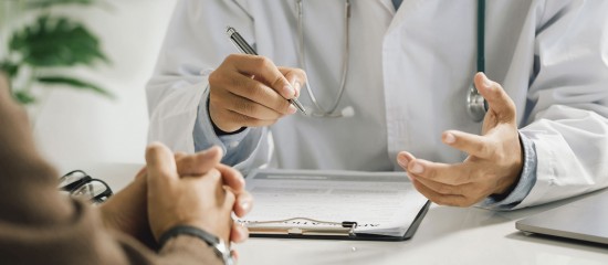 Doctor in a white coat consults with a patient, gesturing while writing on a clipboard.