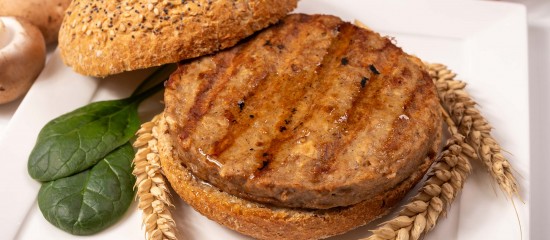 k4_24742652 Grilled beef patty on a toasted sesame bun with spinach leaves and wheat stalks on a white plate.