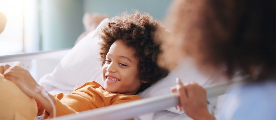 k4_24743138 Smiling child in an orange shirt lying in a hospital bed, connected to an IV line, with a caregiver beside the bed.
