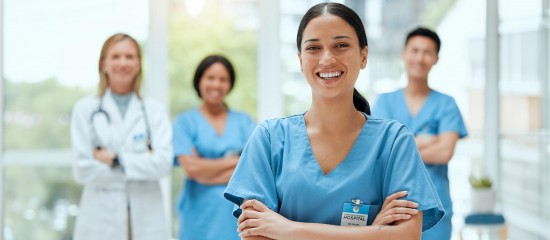 Smiling nurse in blue scrubs with arms crossed, badge visible, standing in front of three colleagues in a bright hospital setting.