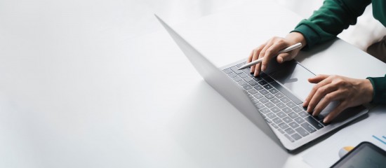 Person typing on a silver laptop at a white desk, wearing a green sweater/sleeve.