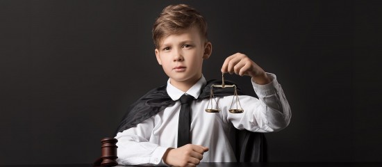 Young boy dressed as a judge, holding scales of justice with a gavel on the bench in front of him.