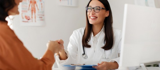 Female doctor with stethoscope shakes hands with a patient at a clinic desk, smiling.
