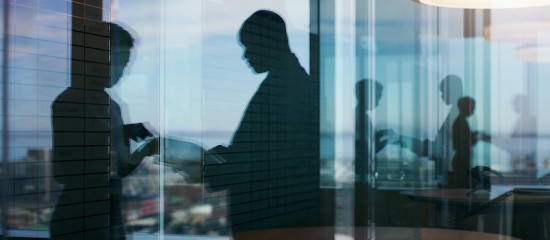Silhouettes of people working behind glass walls in a modern office, with city reflections outside the windows.