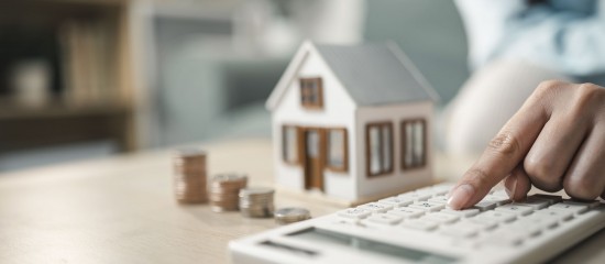 Person uses a calculator to estimate mortgage costs beside a model house and coin stacks.