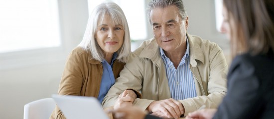 Older couple shakes hands with a professional across a table, a laptop open between them in a bright office environment.