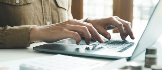 Person typing on a laptop keyboard at a white desk with a blurred notebook nearby.