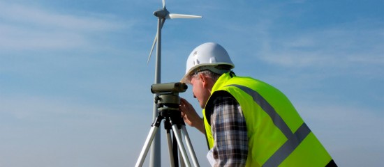 Worker in a high-visibility vest and hard hat uses a surveying instrument on a tripod beside a wind turbine in a blue sky setting.