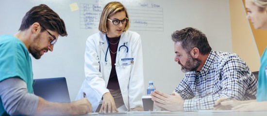 Doctor in a white coat leads a discussion with two colleagues around a table in a clinic setting.
