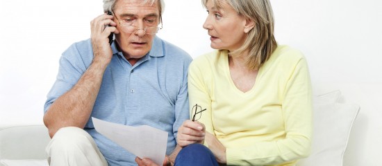 Older couple sitting on a white sofa; man talks on his cellphone while holding papers, woman beside him holding her glasses.