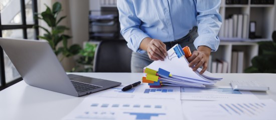 Person organizing reports with color-coded sticky notes beside an open laptop at a white desk.