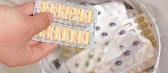Hand holding blister packs of yellow capsules; several more blister packs in a white container in the background.