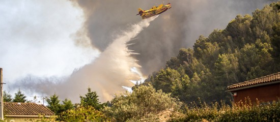 k4_24797438 Yellow air tanker releasing water over a forest fire, with smoke billowing and a hillside covered in trees in the background, and some houses in the foreground.