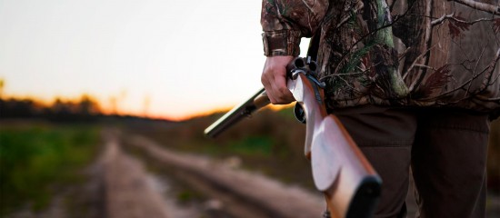 Hunter in camouflage holding a shotgun along a dirt road at sunset.