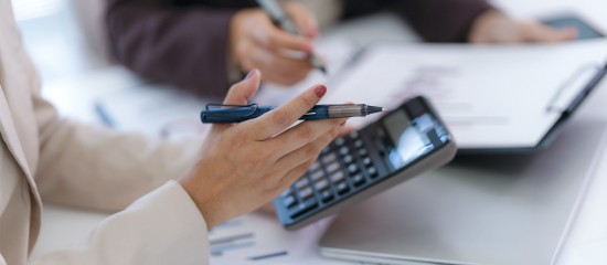 Two professionals review documents with pens, a calculator, and a tablet on a desk, mid-work discussion.