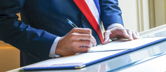 Person in a dark suit signs a document at a desk, with a red, white and blue sash visible.