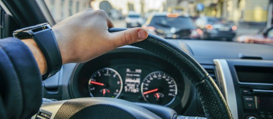 Close-up of a driver’s left hand on the steering wheel with a busy city street outside the car window.