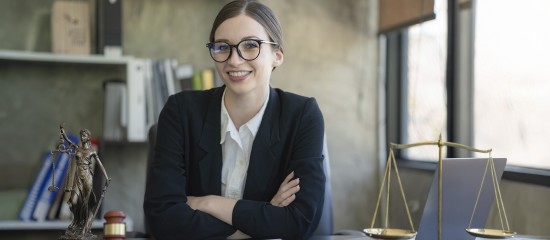 Smiling female lawyer in a black suit sits at a desk with scales of justice in a bright office.