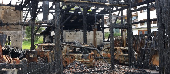 Row of rusted, burned-out cars inside a charred warehouse with blackened beams and rubble scattered around.